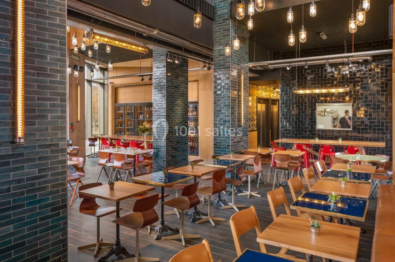 Salle de restaurant moderne avec tables en bois, chaises variées, murs en briques sombres et éclairage suspendu.