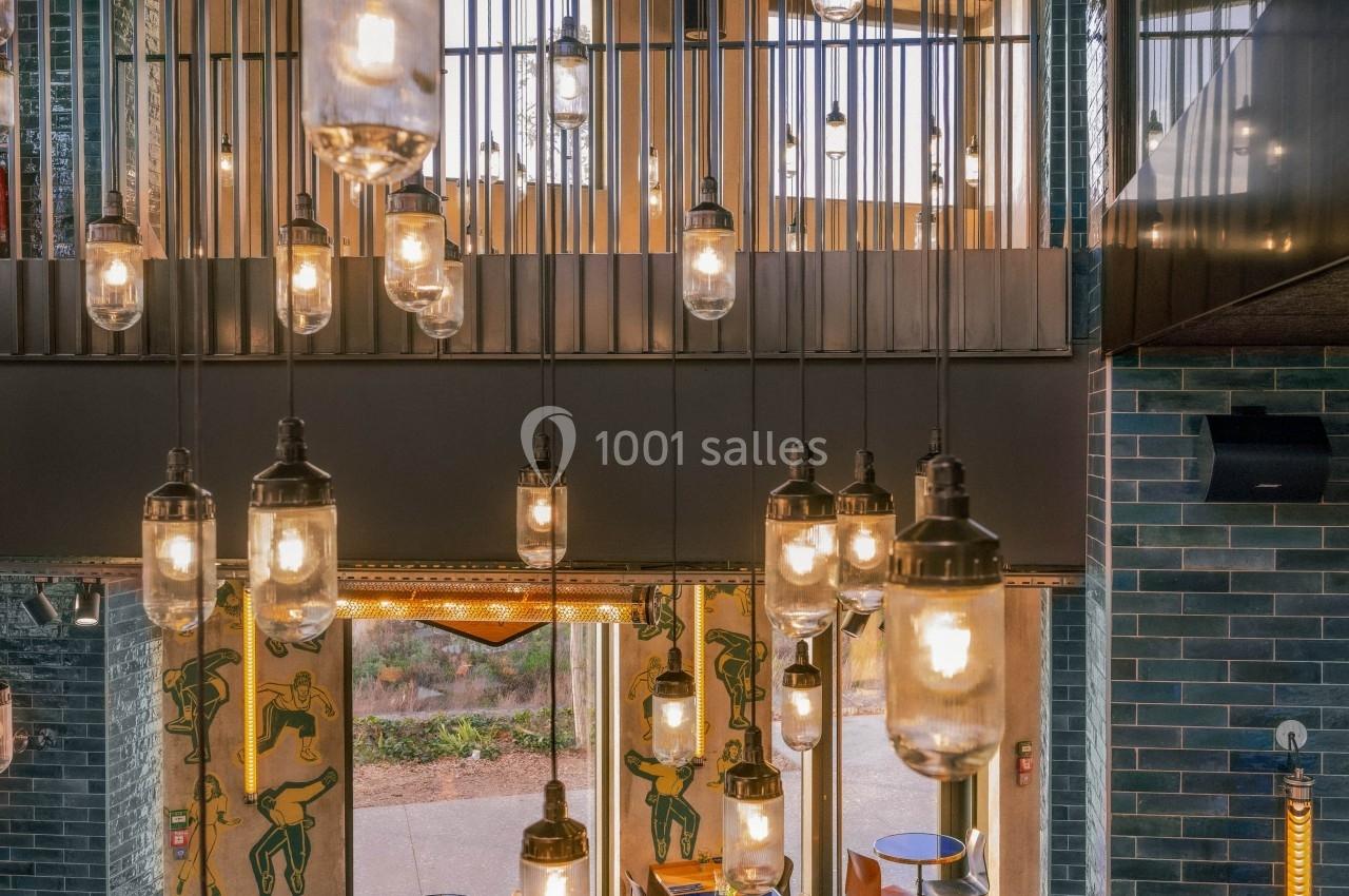 Suspensions lumineuses en verre éclairant un espace intérieur avec tables, chaises orange et murs en briques bleues.