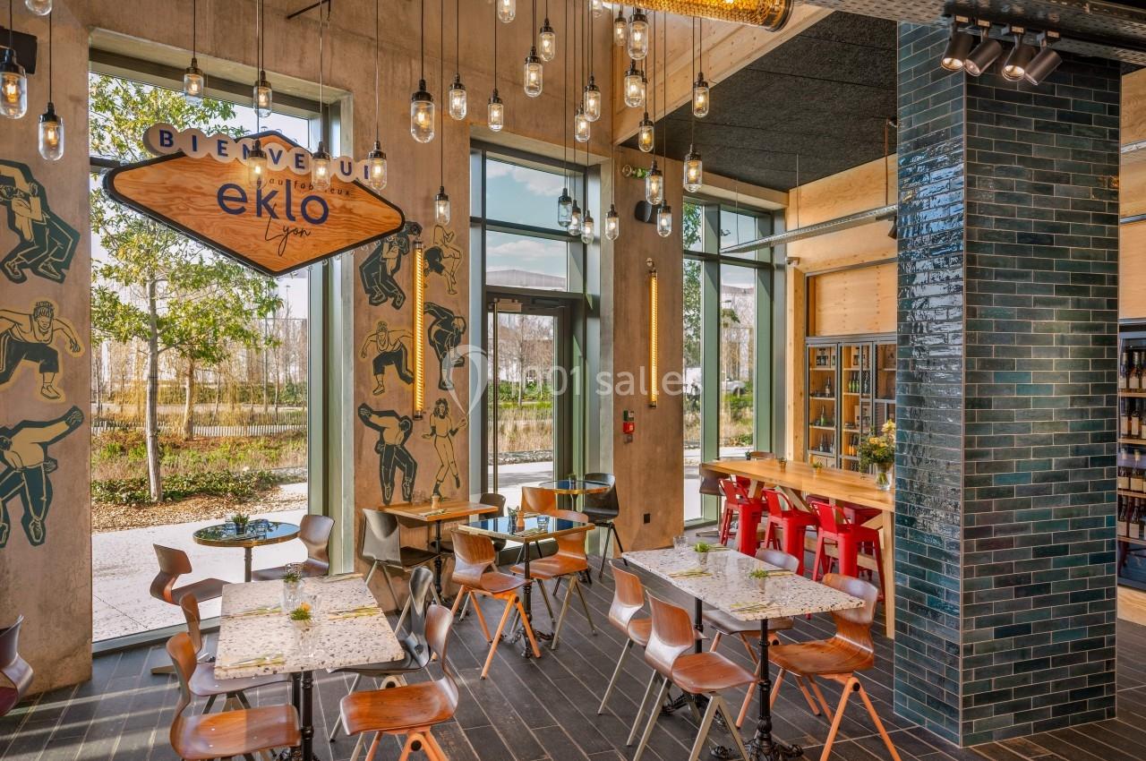 Salle de restaurant lumineuse avec tables en bois, chaises variées, murs décorés et grandes baies vitrées donnant sur l…