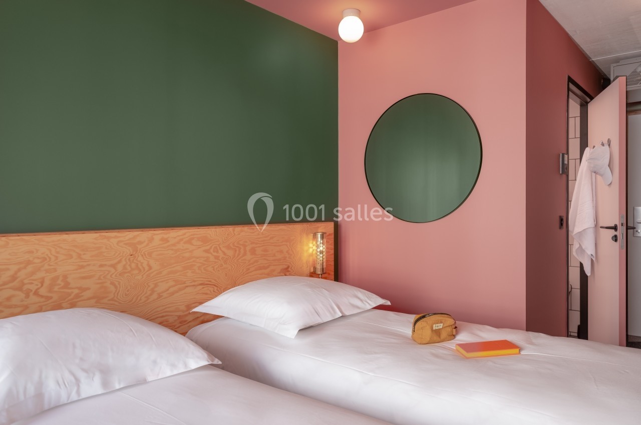 Deux lits simples avec draps blancs dans une chambre aux murs verts et roses, miroir rond et lumière douce.