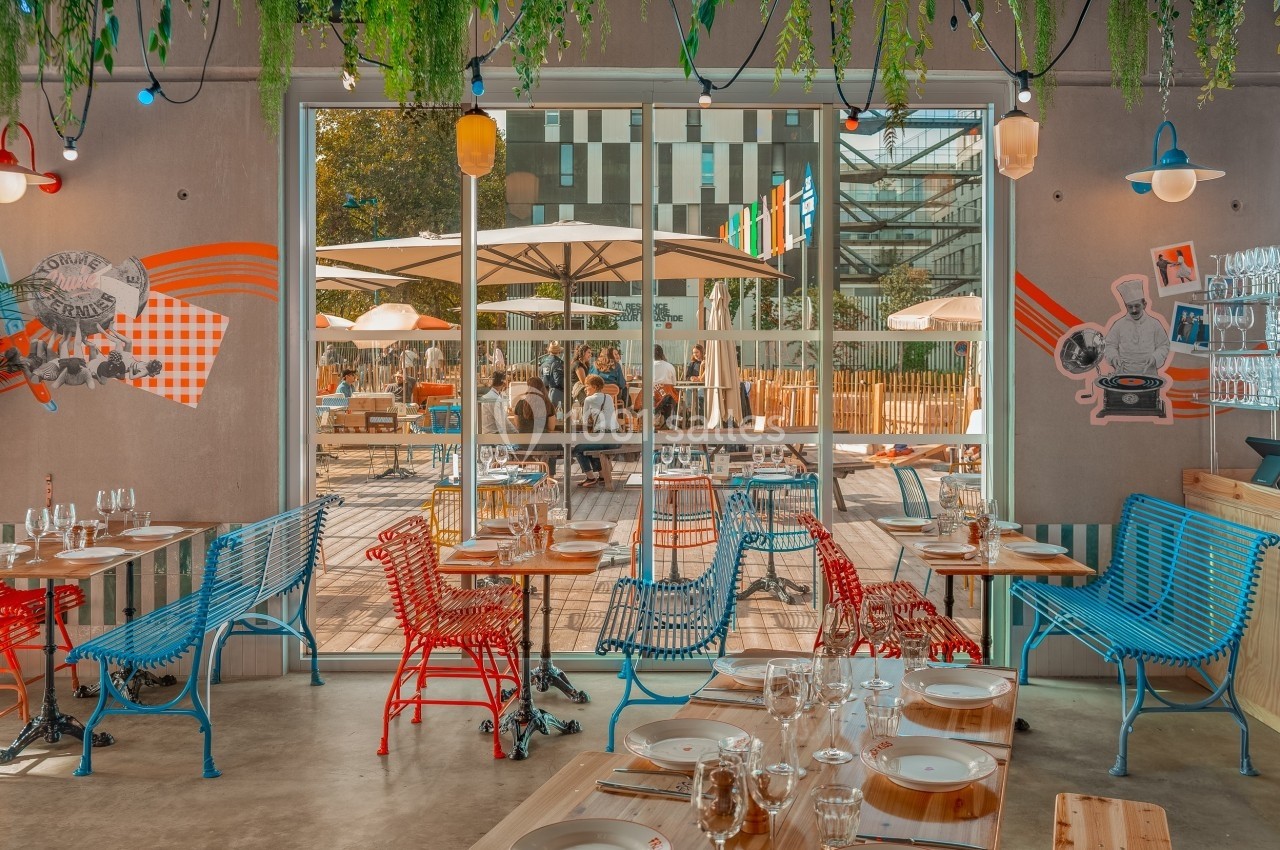 Salle de restaurant colorée avec des tables dressées, donnant sur une terrasse ensoleillée avec des parasols.