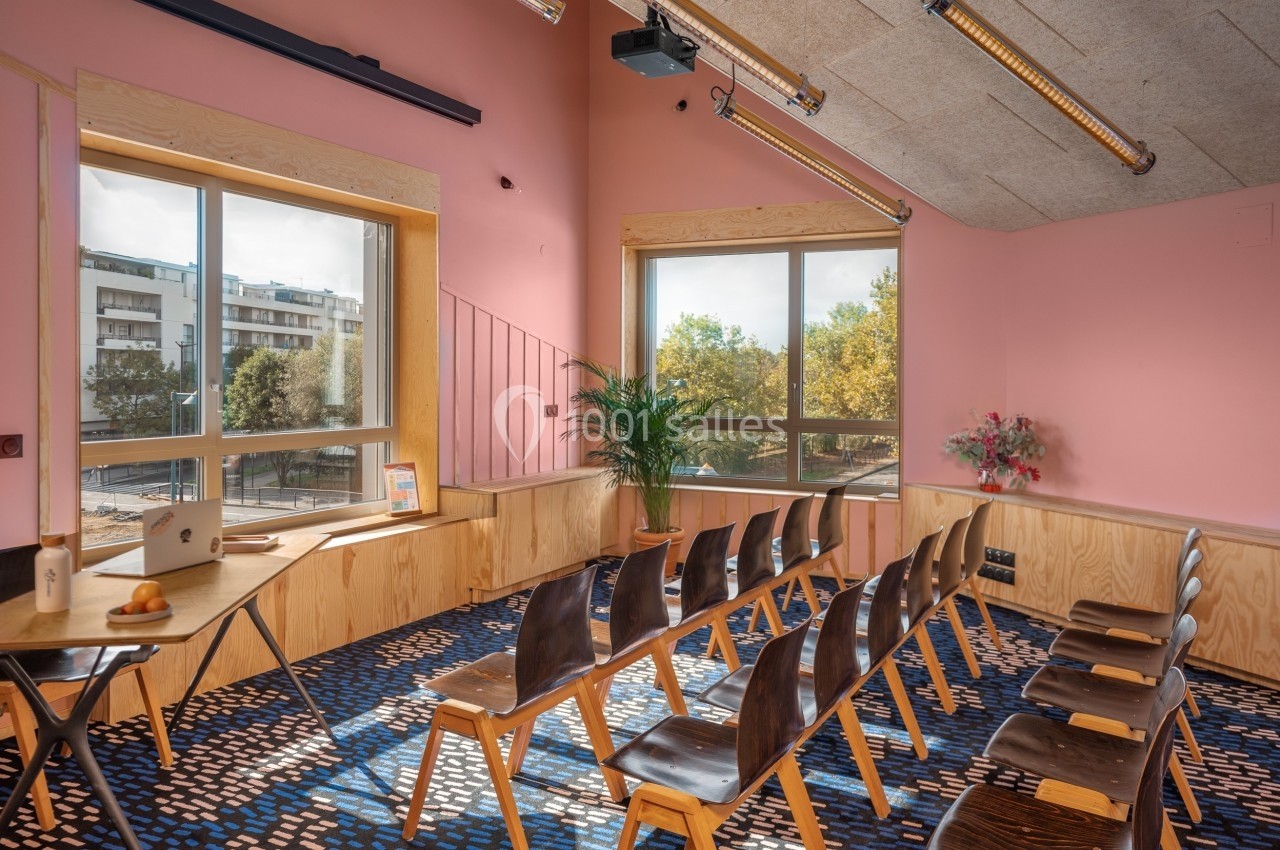Salle lumineuse avec murs roses, grandes fenêtres, chaises alignées, table en bois et plantes décoratives.
