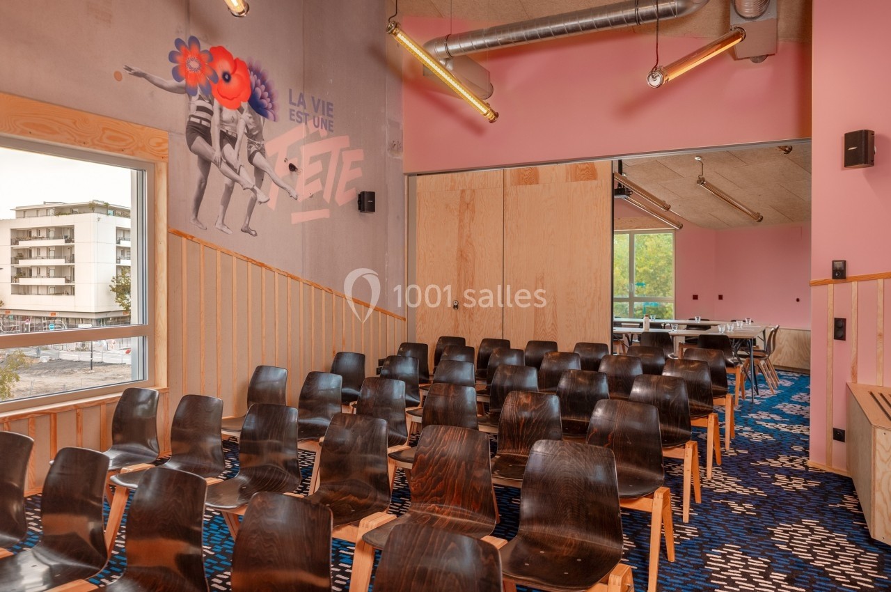 Salle de réunion colorée avec des chaises en bois, mur rose décoré d'une fresque et grande fenêtre donnant sur l'extérieur.