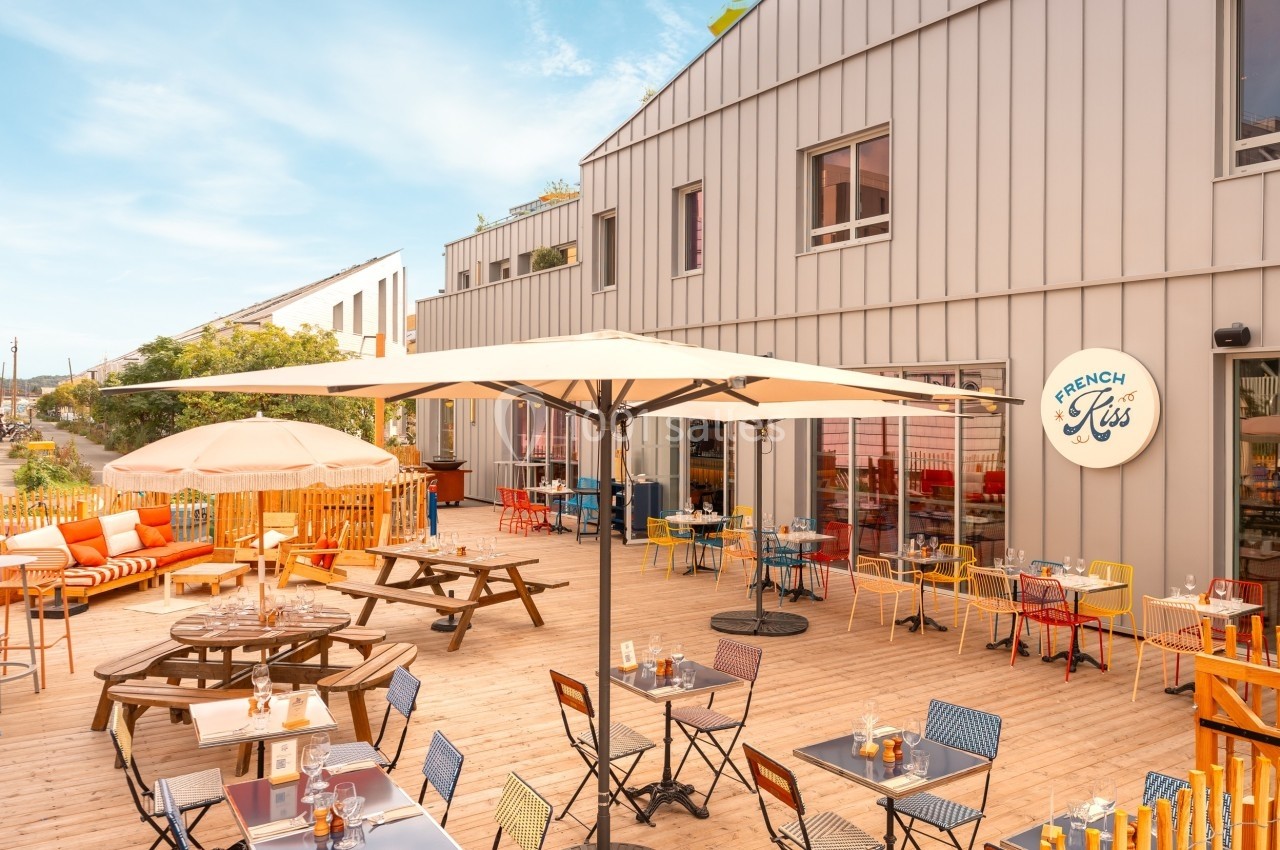 Terrasse en bois avec tables, chaises colorées, parasols et façade d'un bâtiment moderne en arrière-plan.