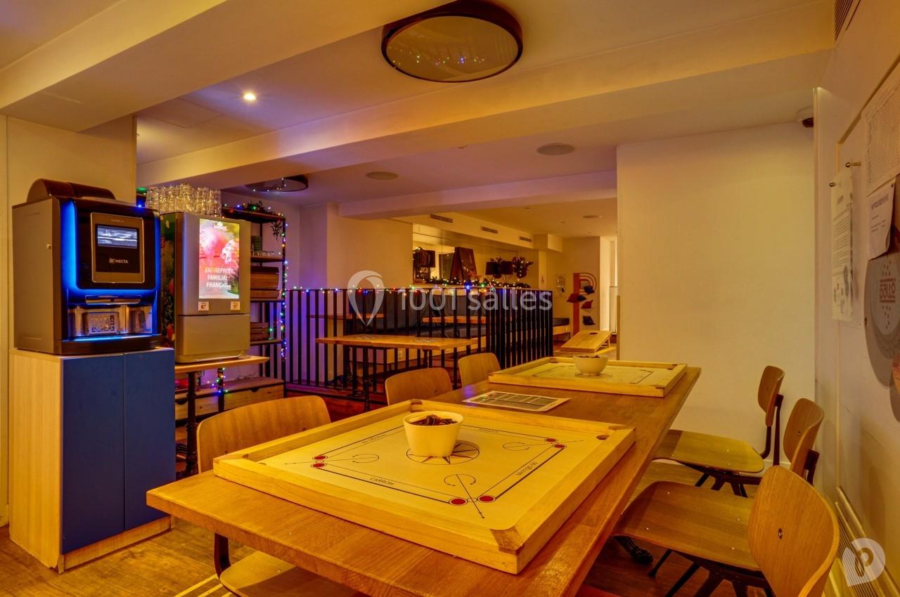 Salle conviviale avec tables en bois, jeux de société, machine à boissons et décoration lumineuse.