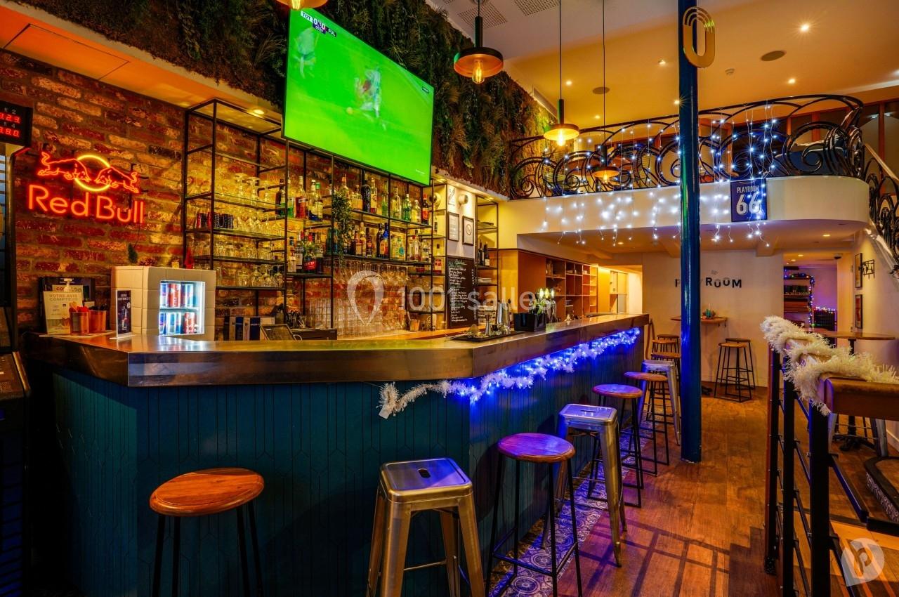 Bar avec comptoir en bois, tabourets métalliques, décorations lumineuses et écran diffusant un match de football.