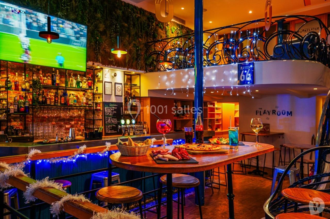 Bar chaleureux avec comptoir en bois, planches apéritives, cocktails, écran diffusant un match et décorations lumineuses.