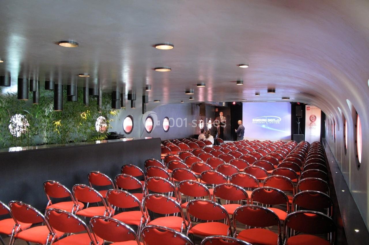 Salle de conférence moderne avec rangées de chaises rouges, éclairage tamisé et écran de présentation à l'arrière.