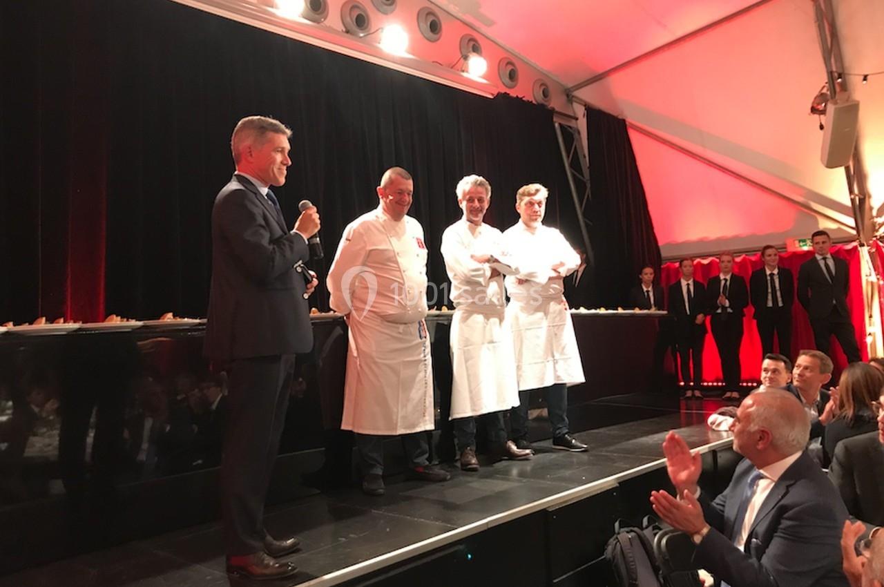 Un homme en costume parle au micro devant trois chefs en tenue blanche sur une scène, sous une tente éclairée.