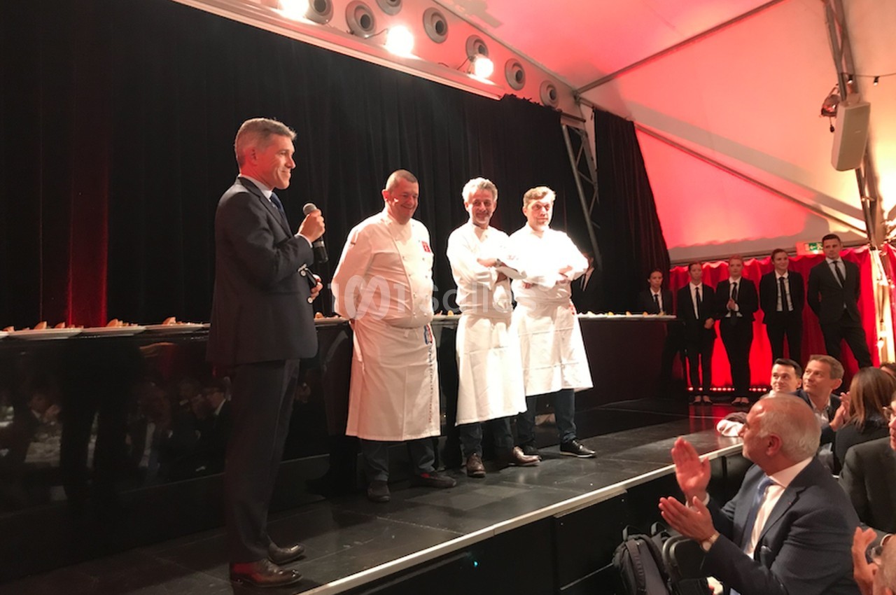 Un homme en costume parle au micro devant trois chefs en tenue blanche sur une scène, sous une tente éclairée.