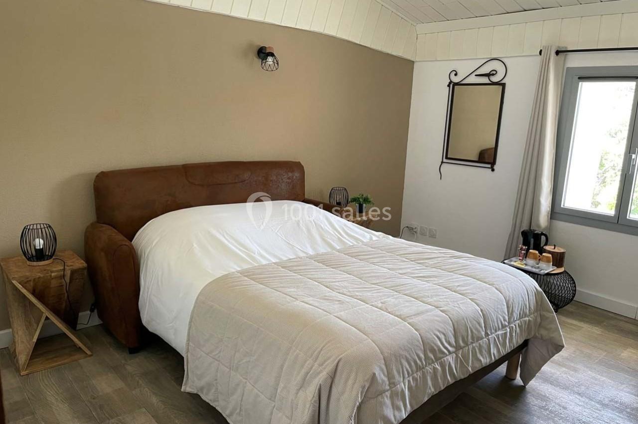 Chambre avec lit double, mur beige, miroir, table de chevet en bois et fenêtre laissant entrer la lumière naturelle.