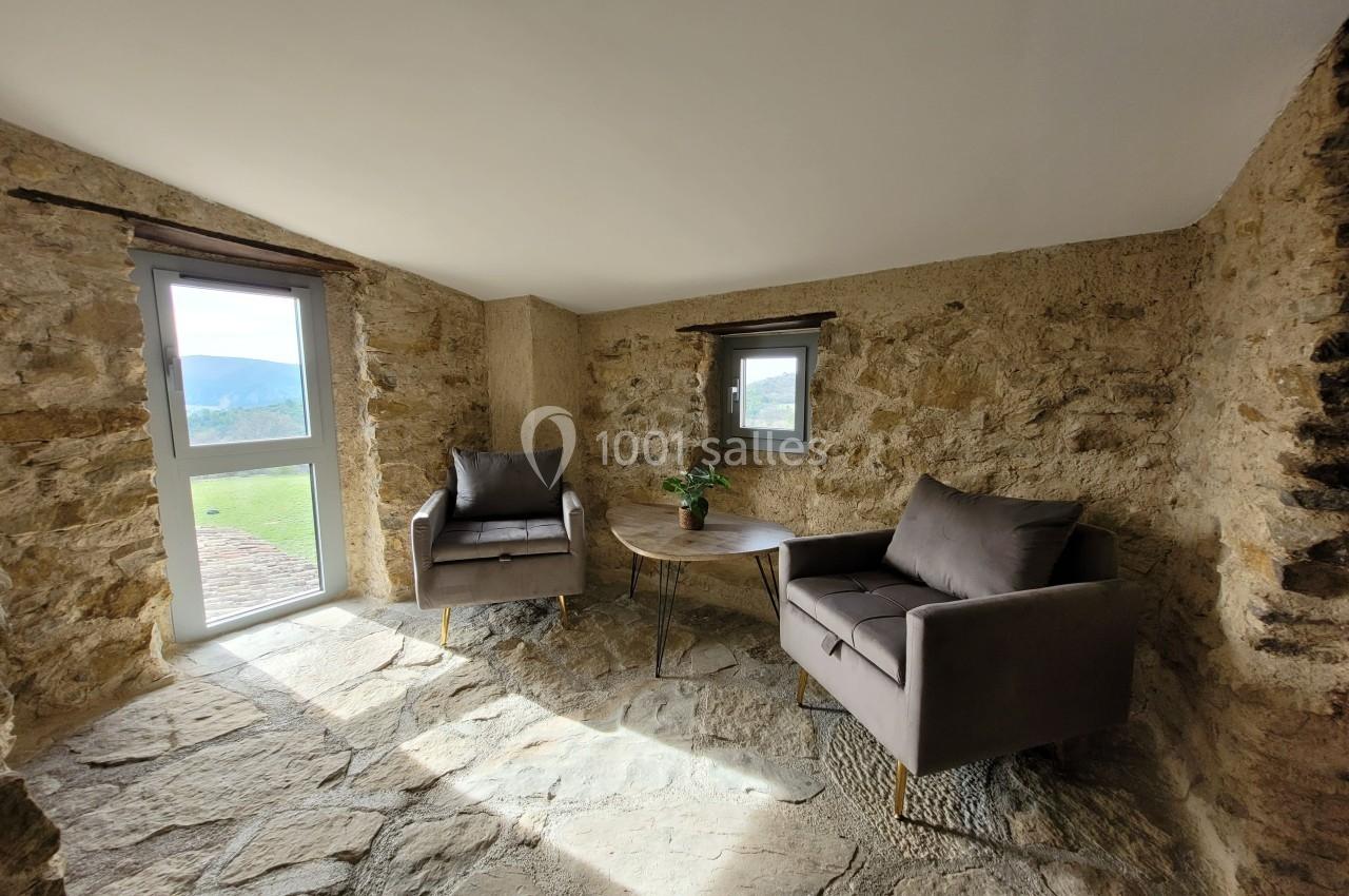 Deux fauteuils gris et une table basse en bois dans une pièce aux murs en pierre avec lumière naturelle.