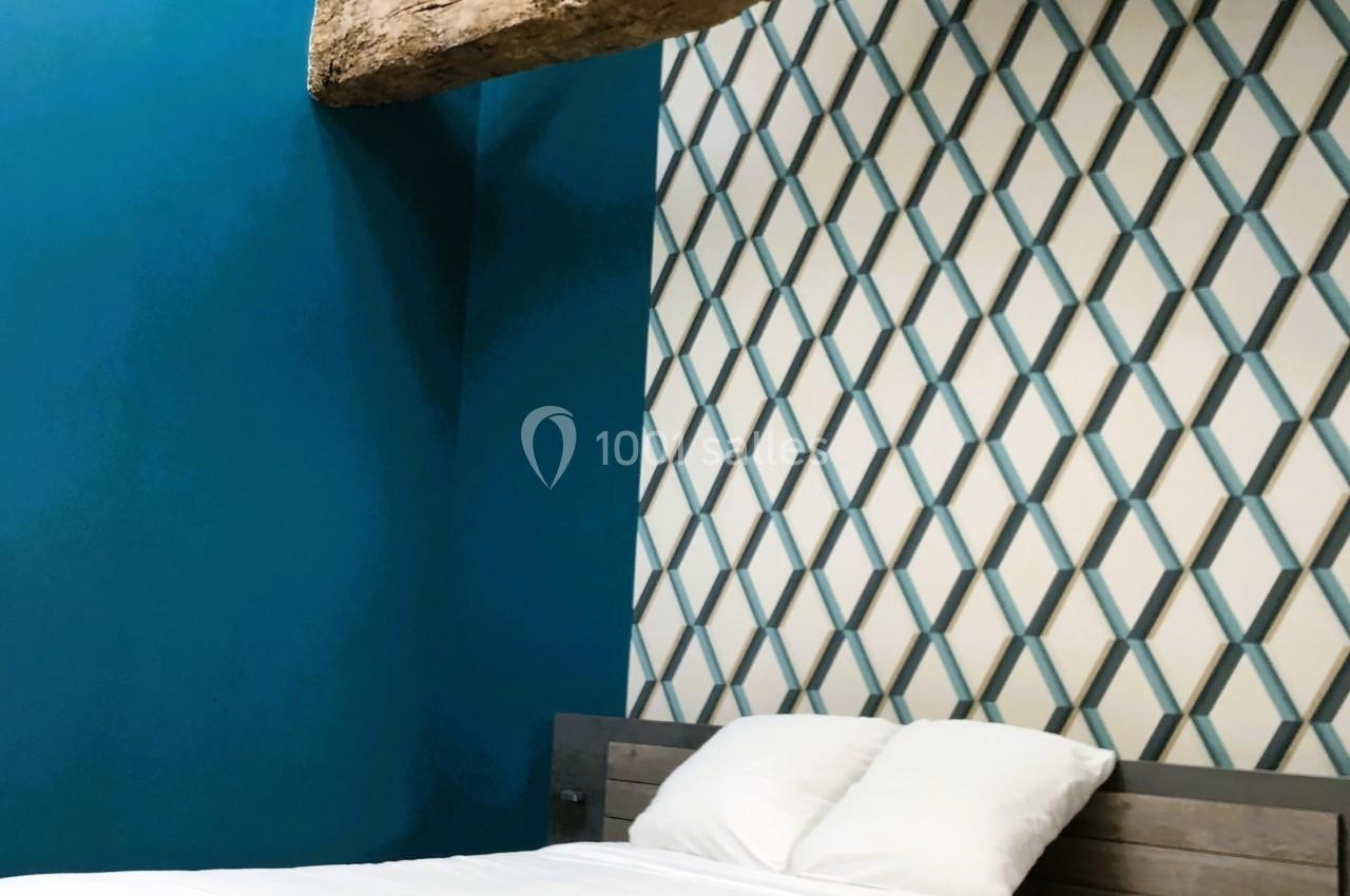 Chambre avec un mur bleu, poutre apparente en bois, lit double en bois clair et mur décoré de motifs géométriques.