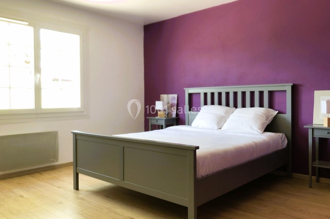 Chambre lumineuse avec un lit double en bois gris, murs blancs et violets, parquet clair et fenêtre à gauche.