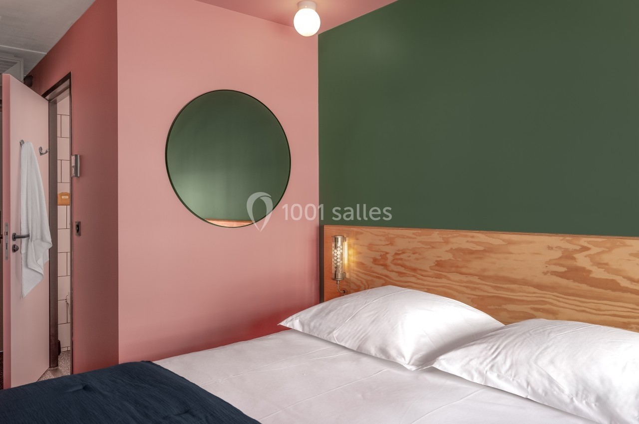 Chambre avec lit double, mur rose et vert, miroir rond, et salle de bain visible par une porte ouverte.