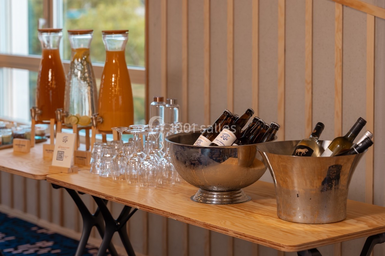 Table avec carafes de jus, verres, bouteilles de bière et de vin dans des seaux en métal, en intérieur.