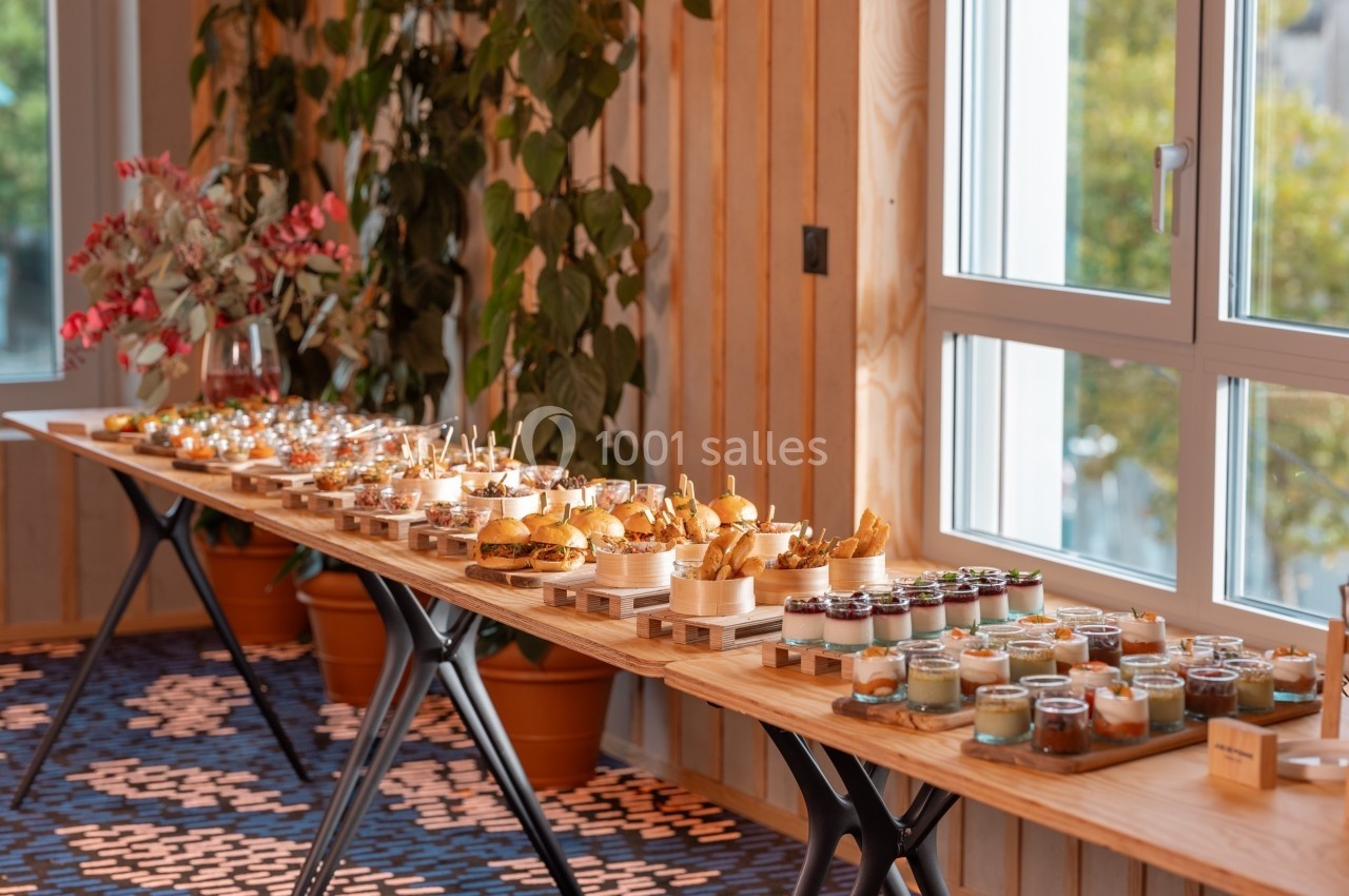 Buffet composé de mini-burgers, verrines et amuse-bouches disposés sur une longue table en bois près d'une fenêtre.