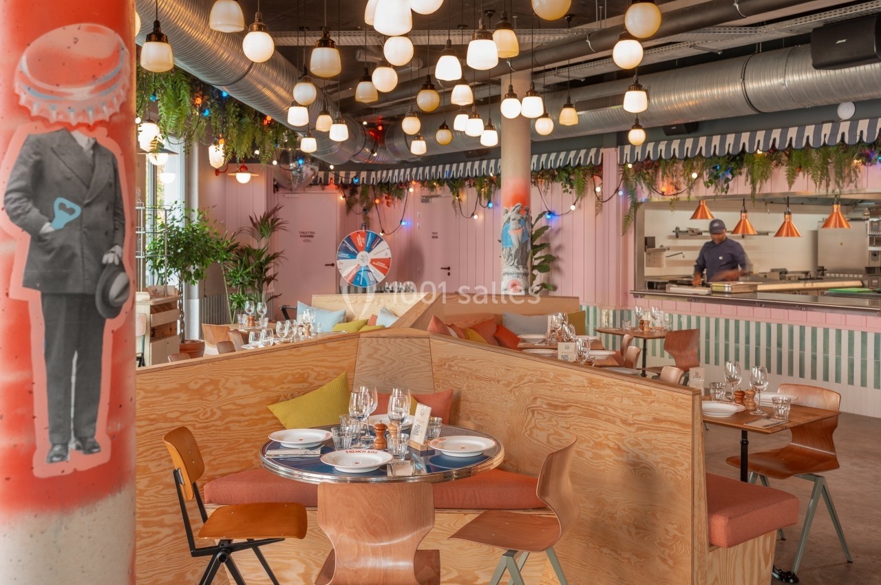 Salle de restaurant moderne avec tables en bois, décor coloré, luminaires suspendus et cuisine ouverte en arrière-plan.