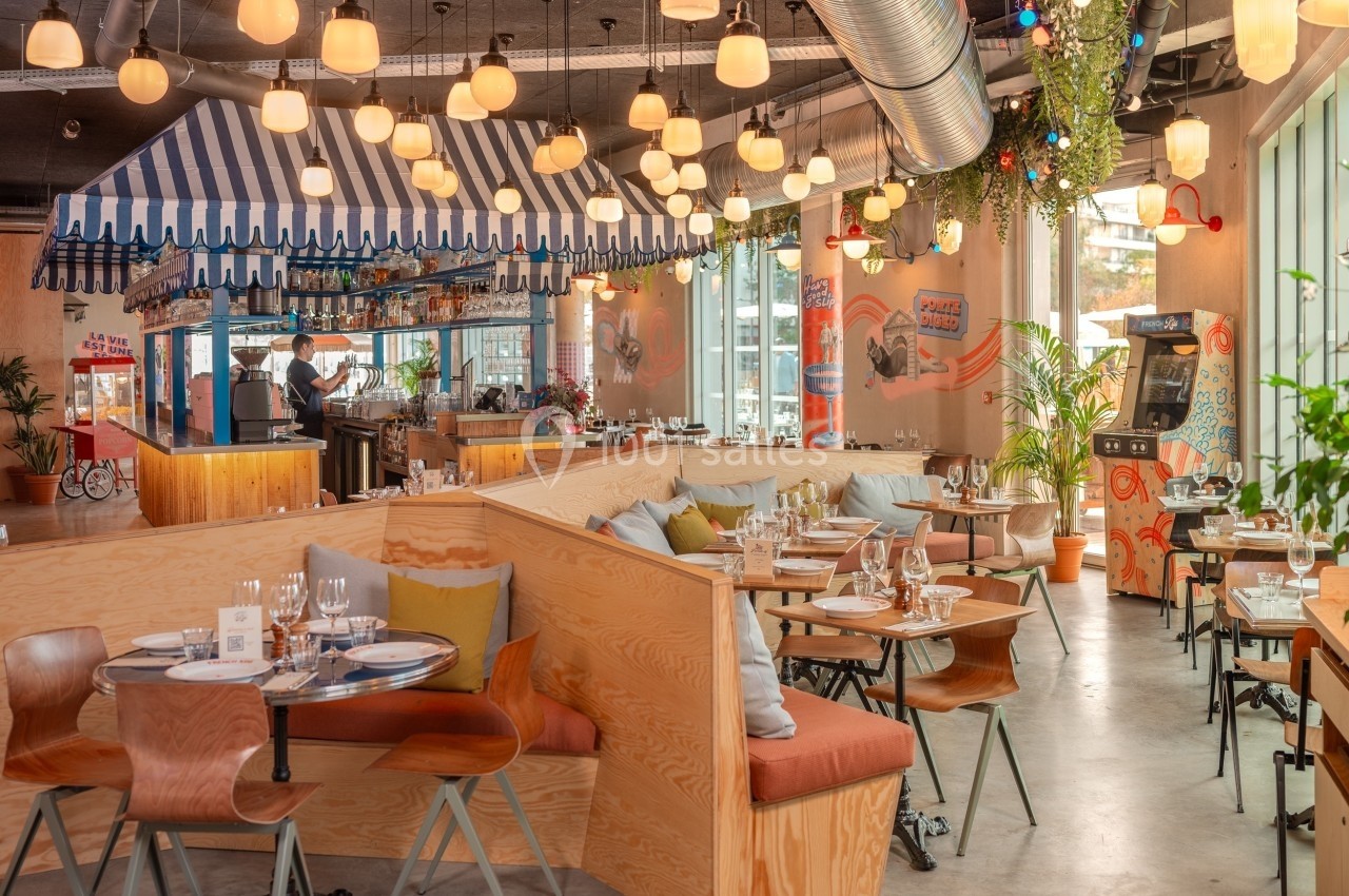 Salle de restaurant lumineuse avec des tables dressées, une décoration colorée et un comptoir sous un auvent rayé.