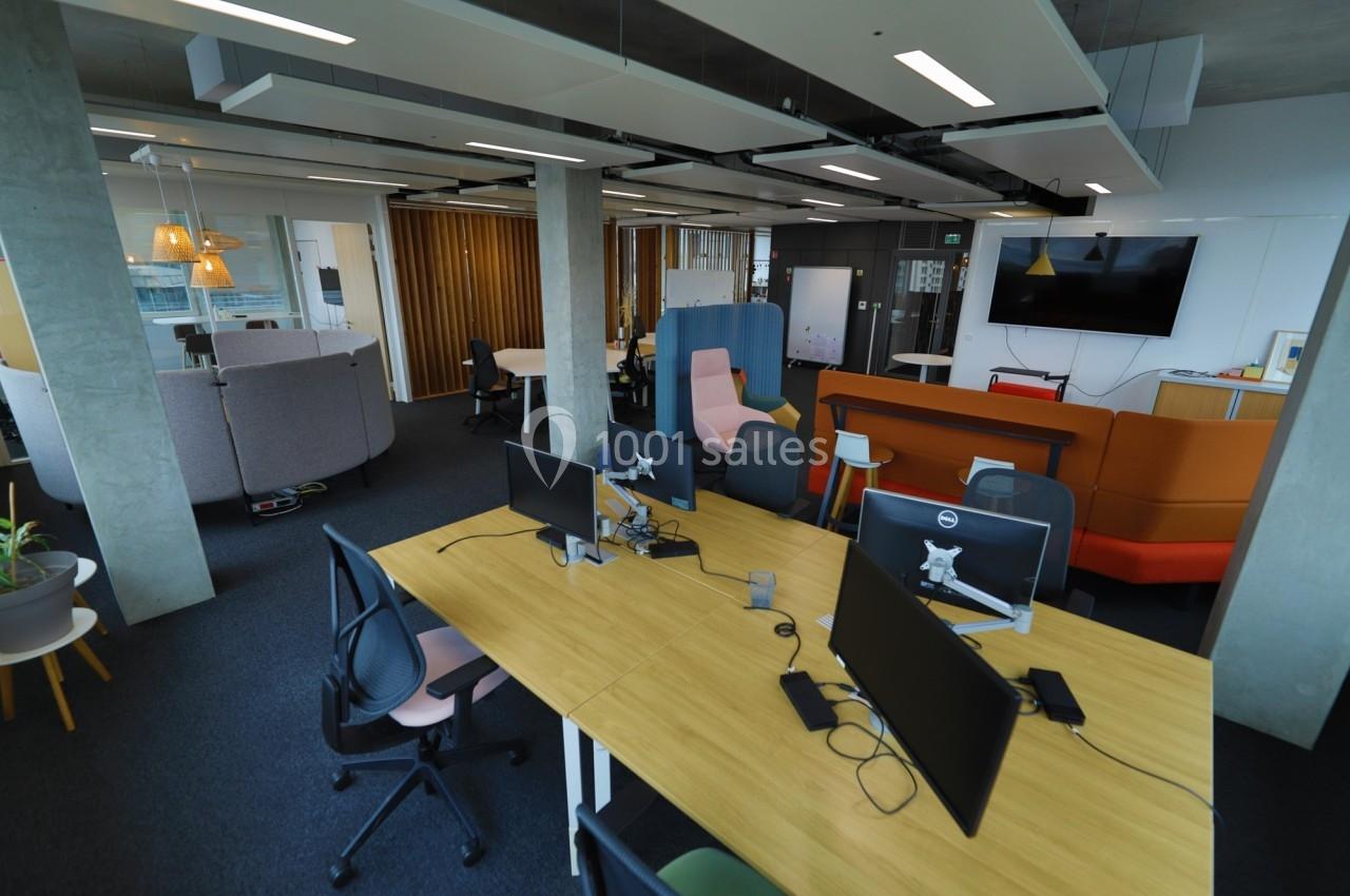 Espace de travail moderne avec bureaux partagés, fauteuils colorés, écran mural et cloisons vitrées.