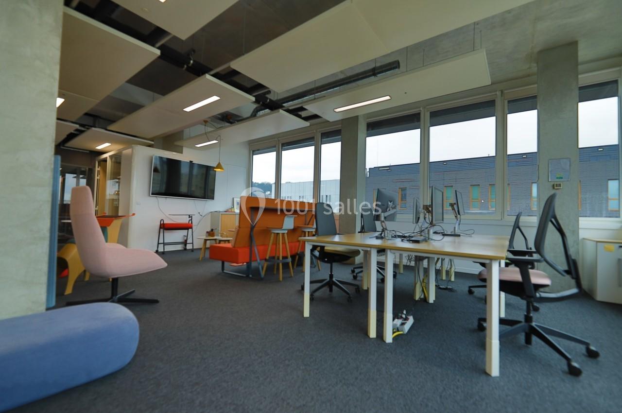 Espace de travail moderne avec bureaux partagés, chaises colorées, grande fenêtre et écran mural.