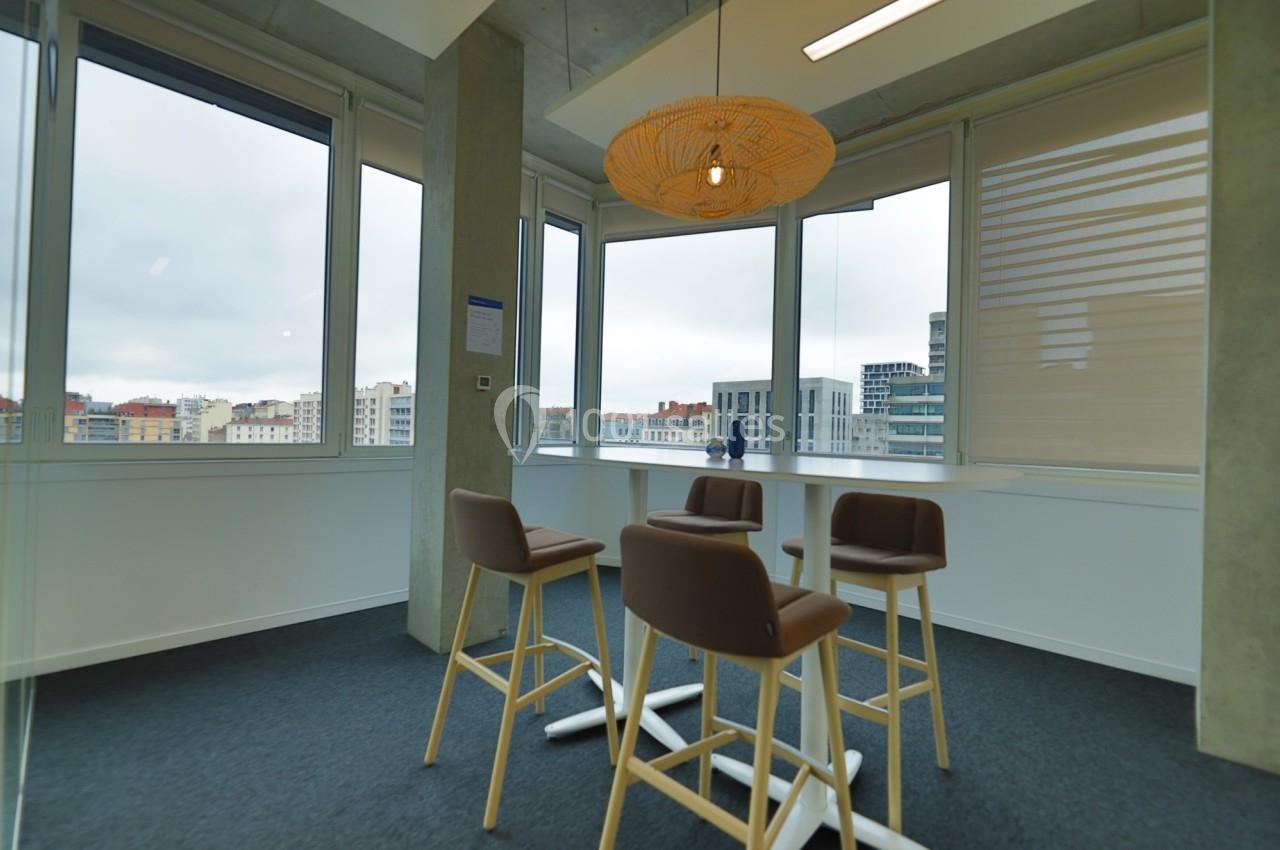 Espace lumineux avec table haute, tabourets et vue sur des bâtiments urbains à travers de grandes fenêtres.