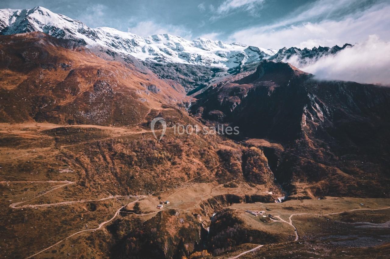 Paysage de montagne avec sommets enneigés, vallées verdoyantes et chemins sinueux sous un ciel partiellement nuageux.