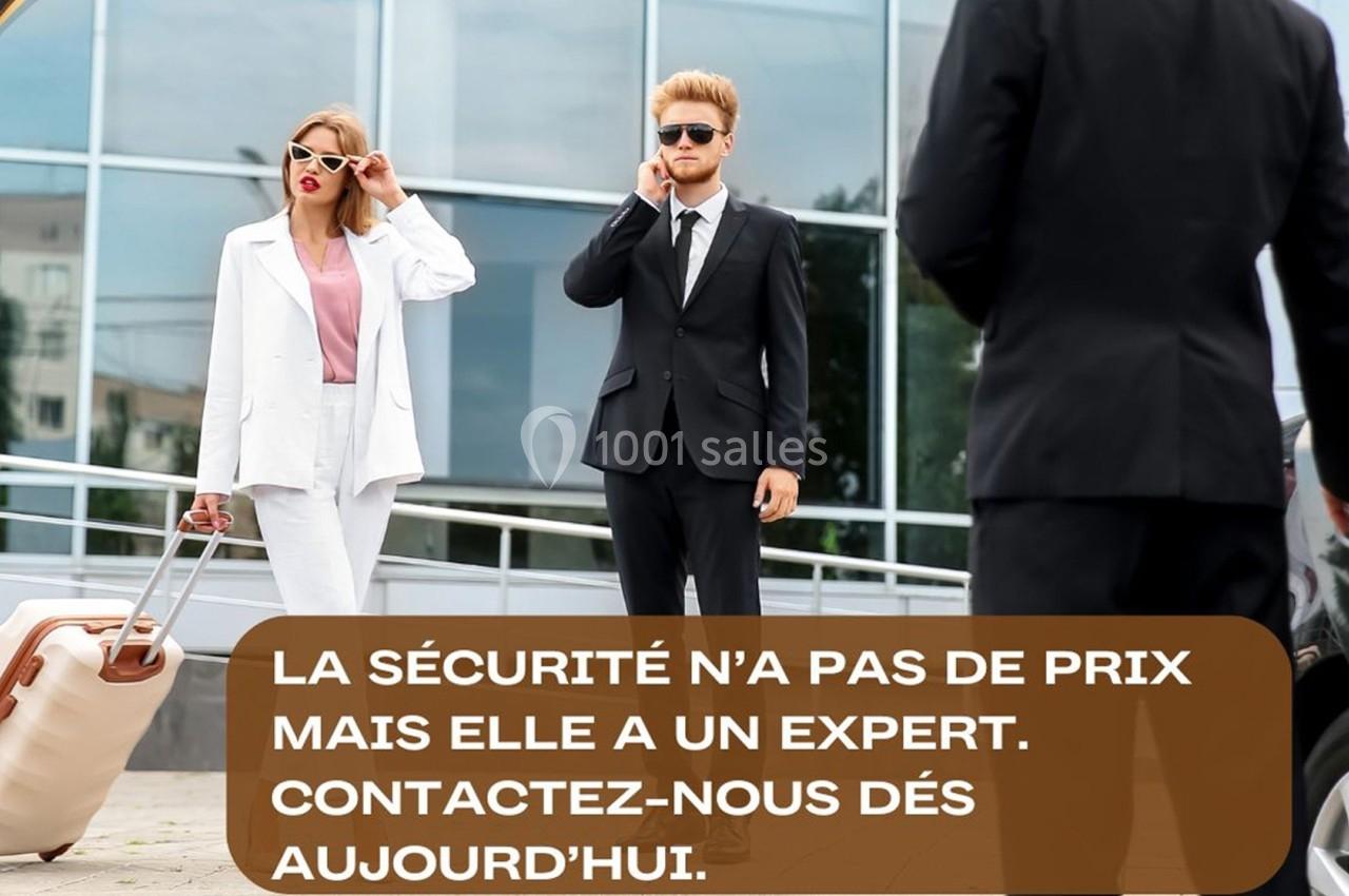 Un homme et une femme en tenue professionnelle avec des valises devant un bâtiment moderne, surveillés par un agent de…
