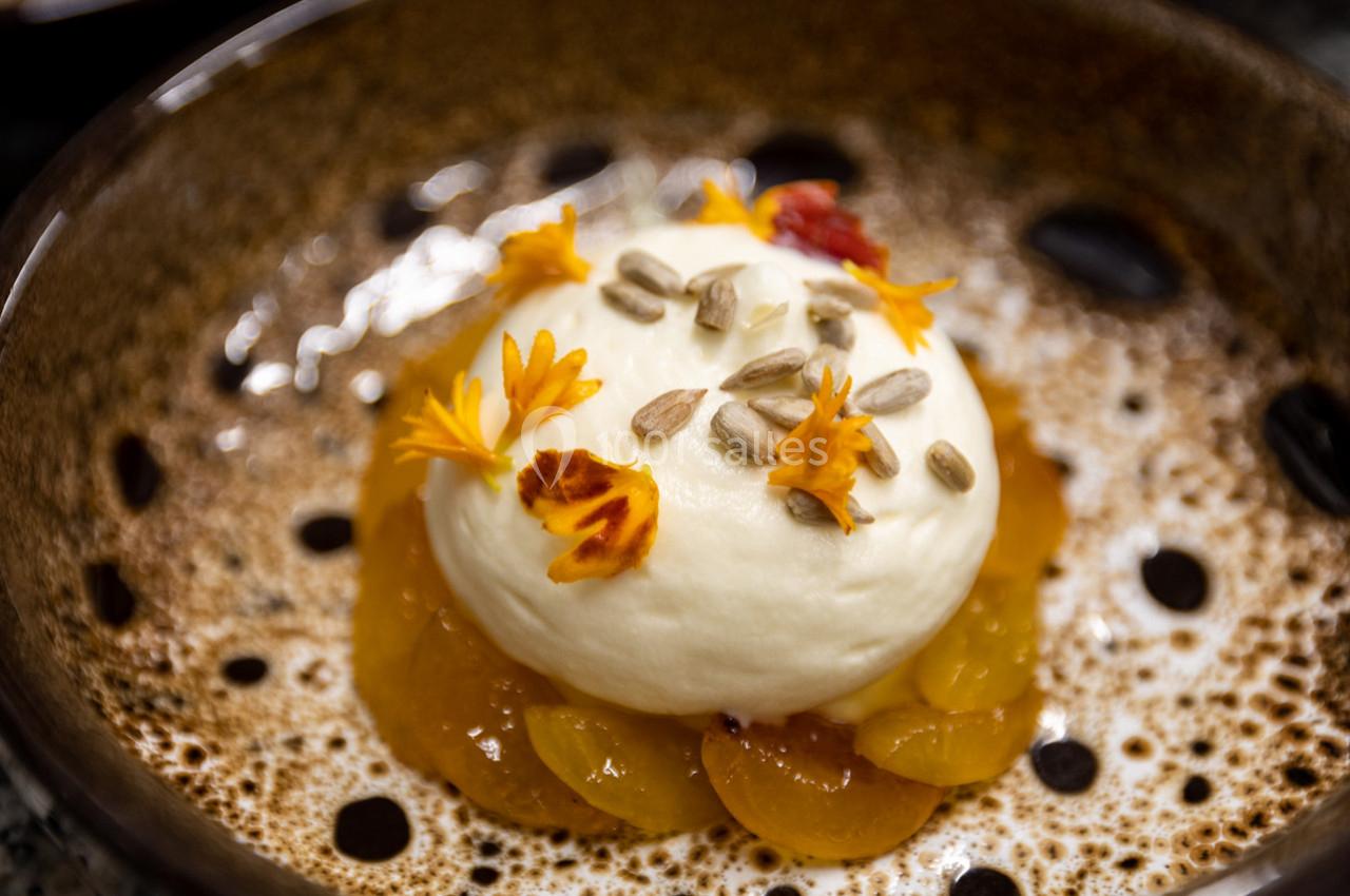 Dessert composé de mousse blanche, pétales de fleurs, graines de tournesol et fruits jaunes dans une assiette brune.