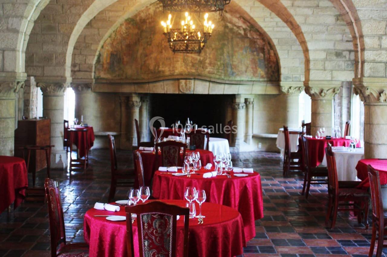 Salle de restaurant élégante avec tables dressées, nappes rouges, chandeliers suspendus et murs en pierre voûtés.