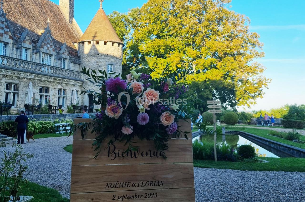 Panneau en bois décoré de fleurs devant un château avec jardin, canal et ciel dégagé en arrière-plan.