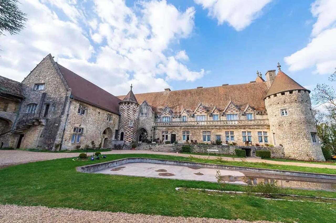 Château en pierre avec tours et toits en tuiles, entouré d'une pelouse et d'une allée gravillonnée, sous un ciel…