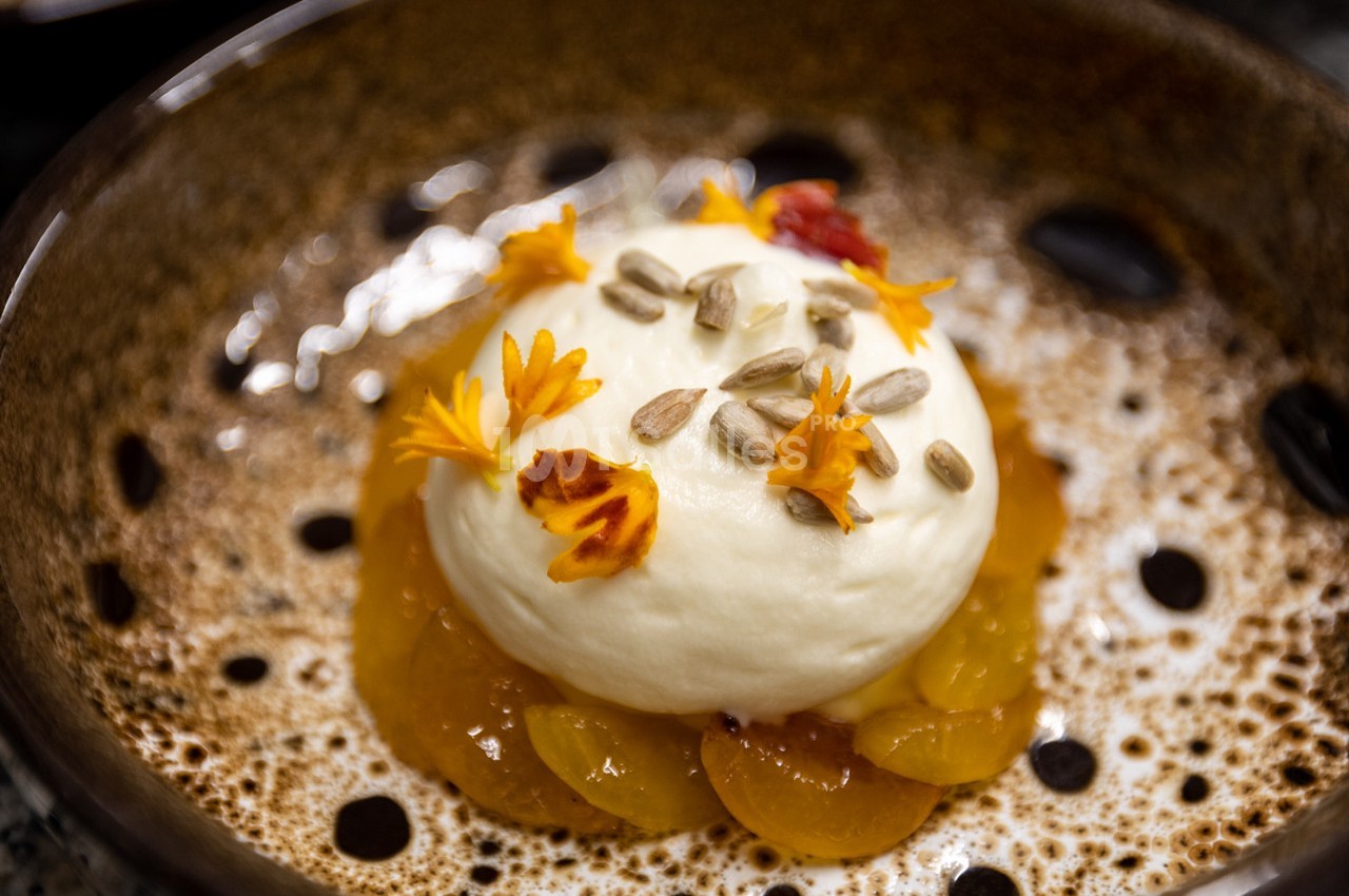 Dessert composé de mousse blanche, pétales de fleurs, graines de tournesol et fruits jaunes dans une assiette brune.