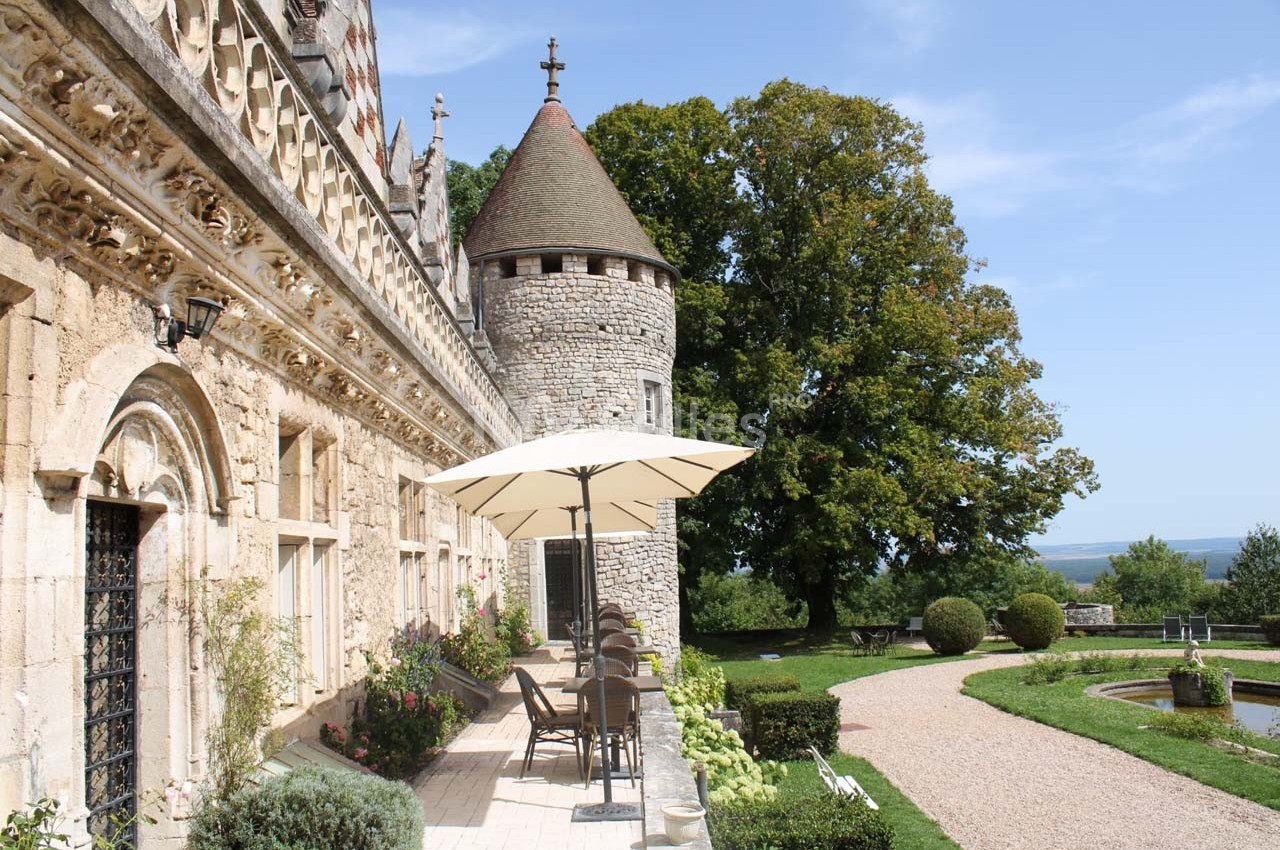 Façade en pierre d'un château avec une tour ronde, terrasse aménagée et jardin verdoyant sous un ciel dégagé.