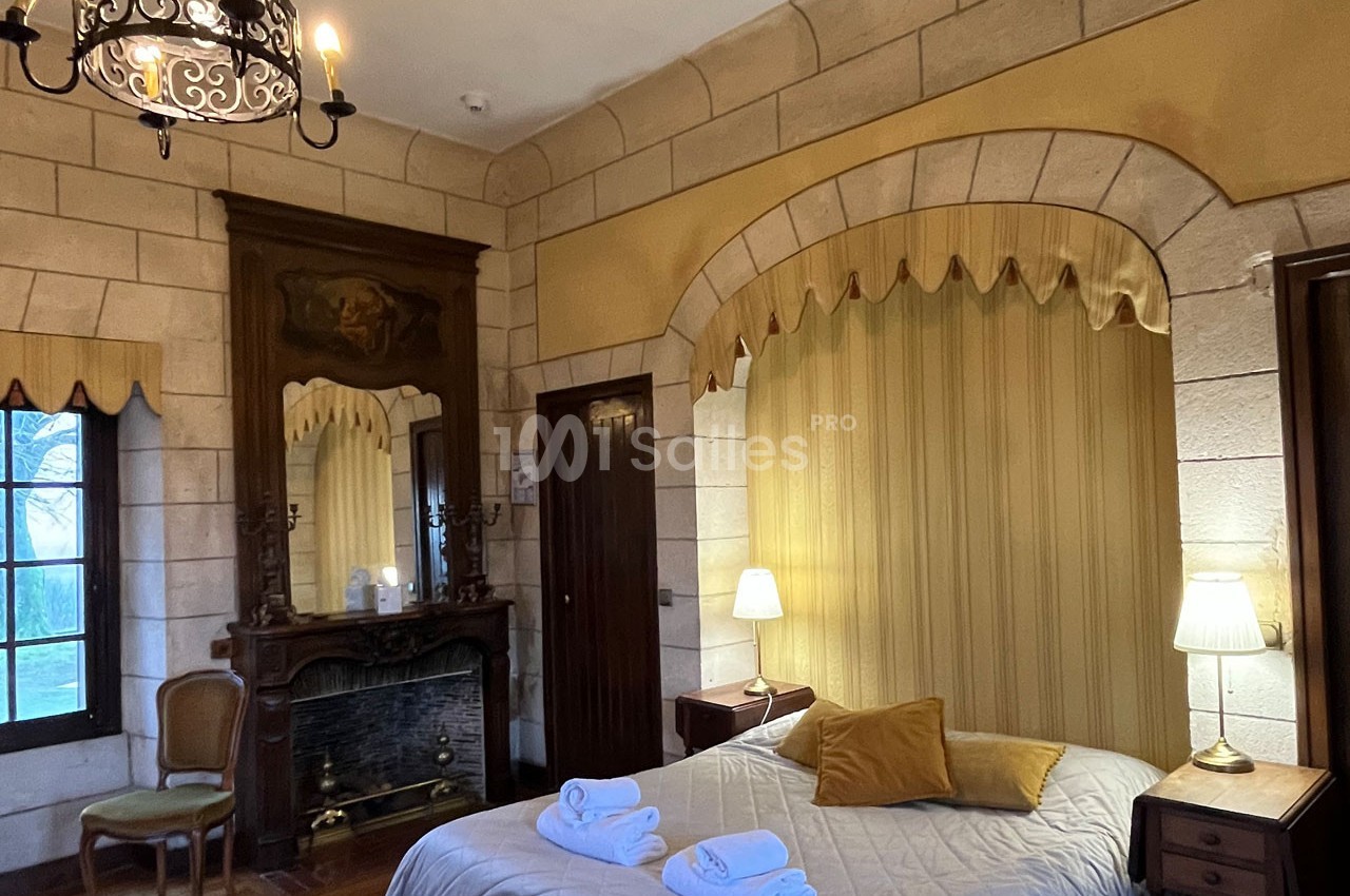 Chambre avec murs en pierre, lit double, cheminée ancienne, miroir, et éclairage doux par des lampes de chevet.