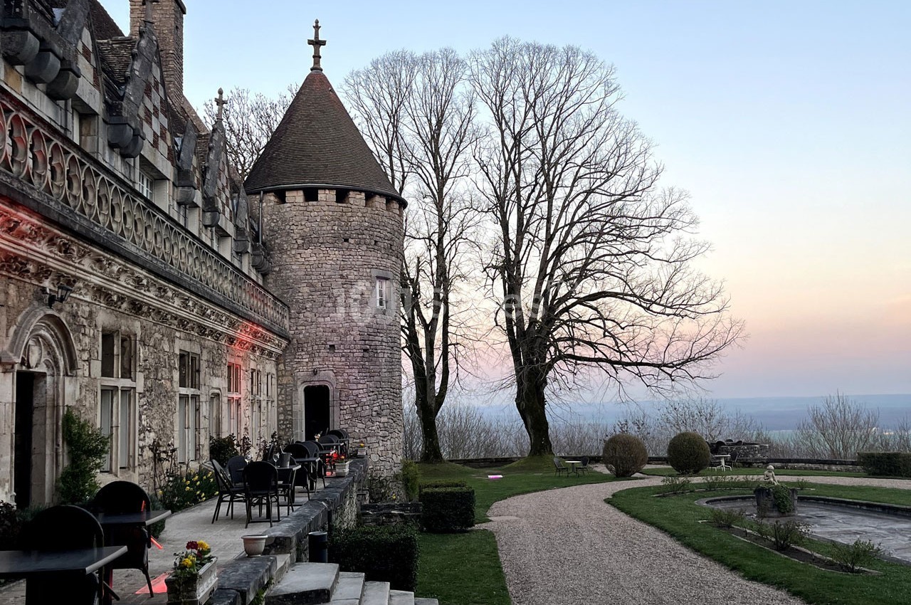 Château en pierre avec une tour ronde, terrasse aménagée et jardin, au crépuscule sous un ciel dégagé.
