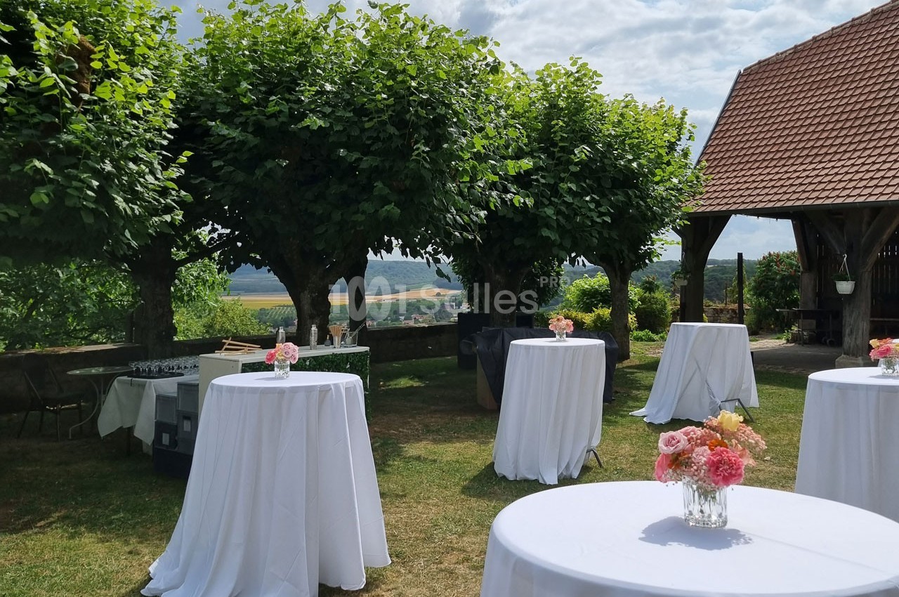 Tables hautes recouvertes de nappes blanches et décorées de fleurs, disposées dans un jardin ombragé.