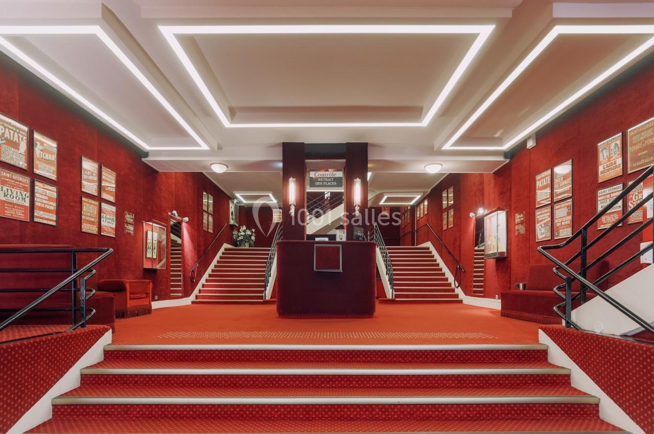 Hall d'entrée d'un théâtre avec tapis rouge, murs ornés d'affiches et escaliers symétriques menant à l'étage.