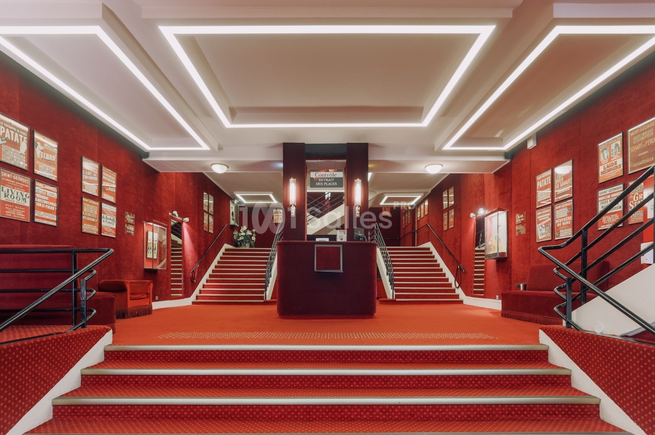 Hall d'entrée d'un théâtre avec tapis rouge, murs ornés d'affiches et escaliers symétriques menant à l'étage.