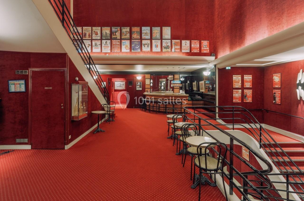 Intérieur d'un théâtre avec moquette rouge, affiches encadrées aux murs, tables et chaises près d'une rampe.