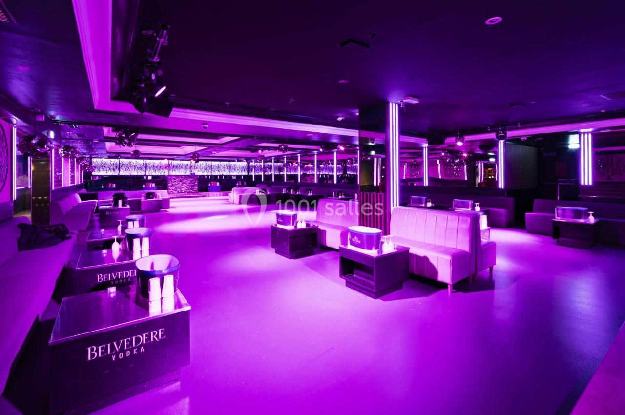 Salle de club nocturne avec éclairage violet, canapés blancs, tables basses et piste de danse dégagée.