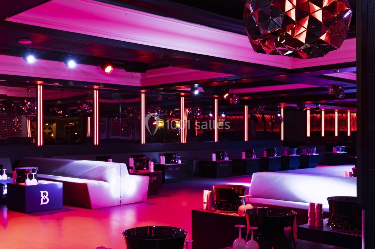 Salle de club avec éclairage rouge et violet, canapés blancs et tables basses équipées de seaux à champagne.