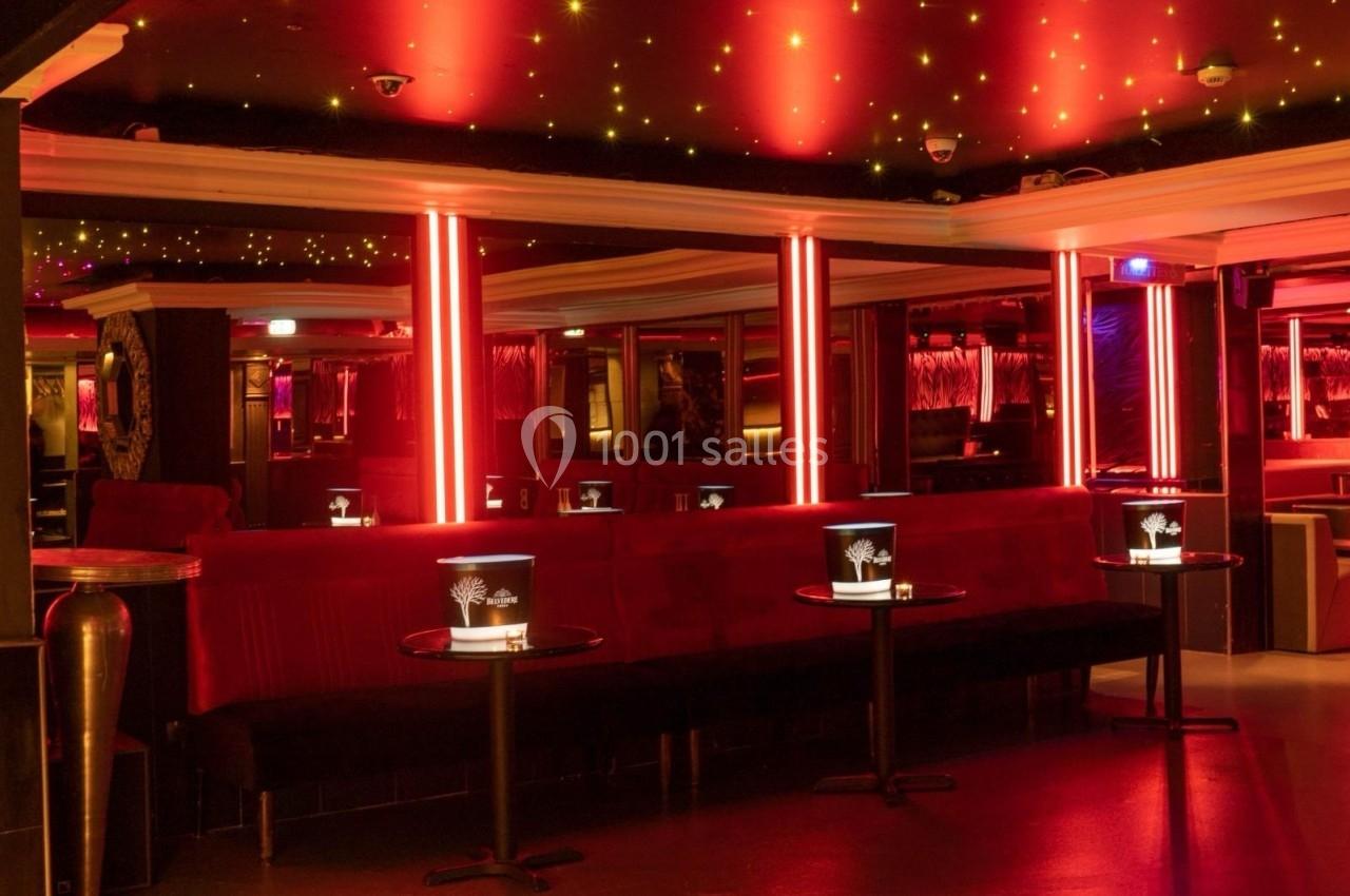 Salle de club avec éclairage rouge, banquettes en velours, tables basses et seaux à champagne.