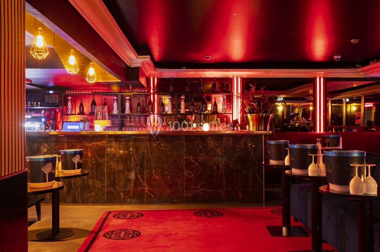Intérieur d'un bar lounge avec éclairage rouge, comptoir en marbre sombre et tables décorées de seaux à champagne.