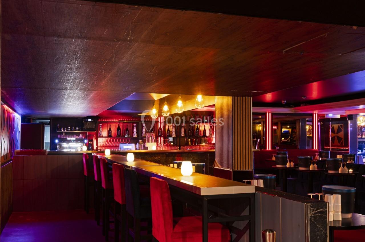 Intérieur d'un bar avec un long comptoir, des tabourets rouges, des lumières tamisées et une ambiance chaleureuse.