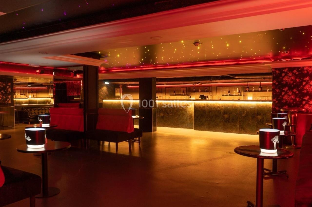 Salle de bar avec éclairage rouge tamisé, tables basses noires et comptoir rétroéclairé en arrière-plan.