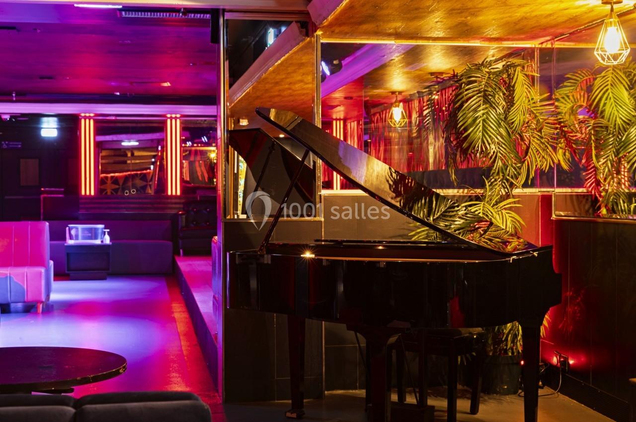 Piano à queue noir dans un intérieur de bar ou club avec éclairage tamisé et décorations végétales.