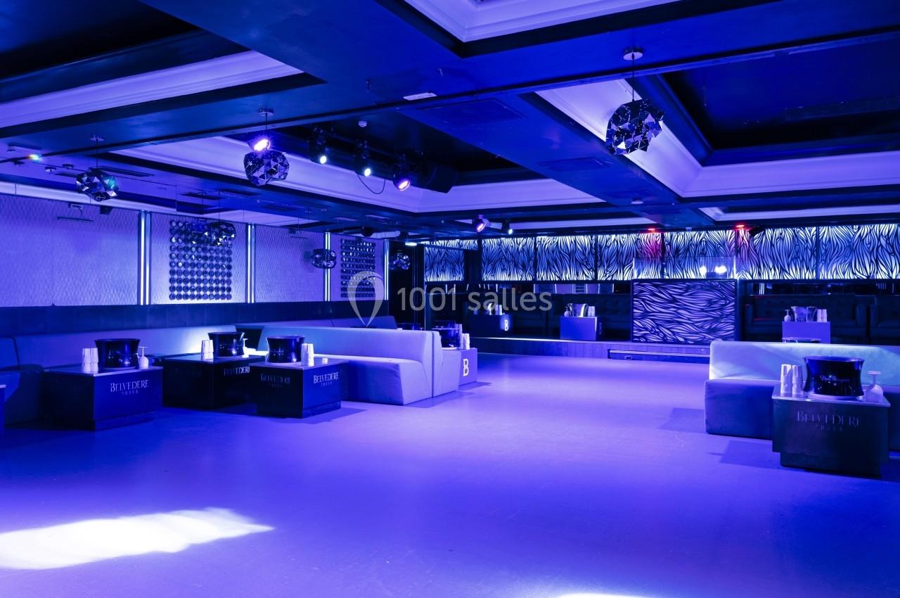 Salle de club nocturne moderne avec éclairage bleu, banquettes, tables basses et décorations murales texturées.