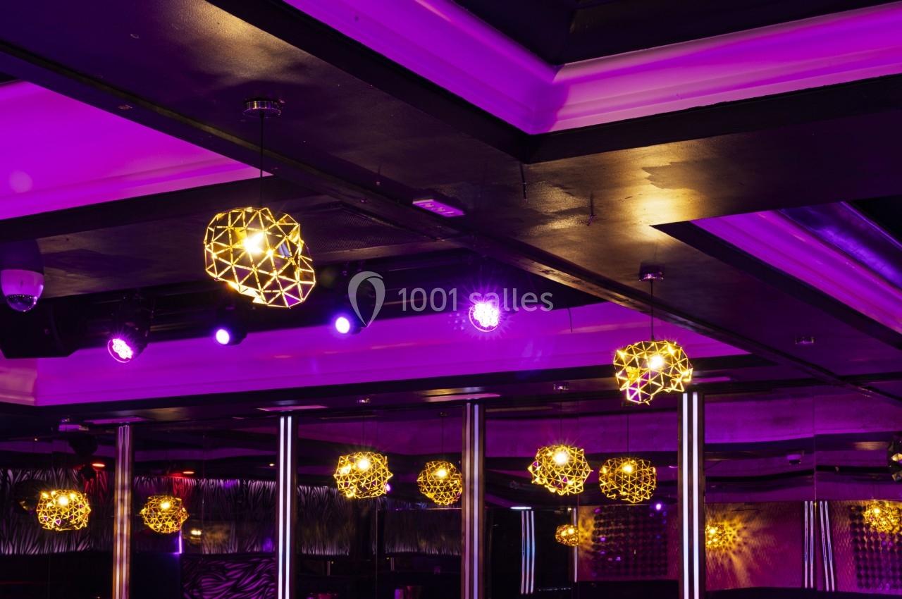 Plafond d'une salle avec des luminaires géométriques dorés suspendus, éclairés par une lumière violette.