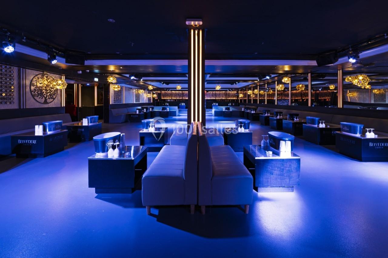 Salle de club moderne avec éclairage bleu, canapés, tables basses et décorations murales élégantes.